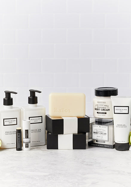 Products from Beekman 1802's Fresh Air collection all displayed together showing the bar soaps, body cream, hand & Body wash, hand cream, lotion and lip balm, surrounded by limes on a light gray tile background.  