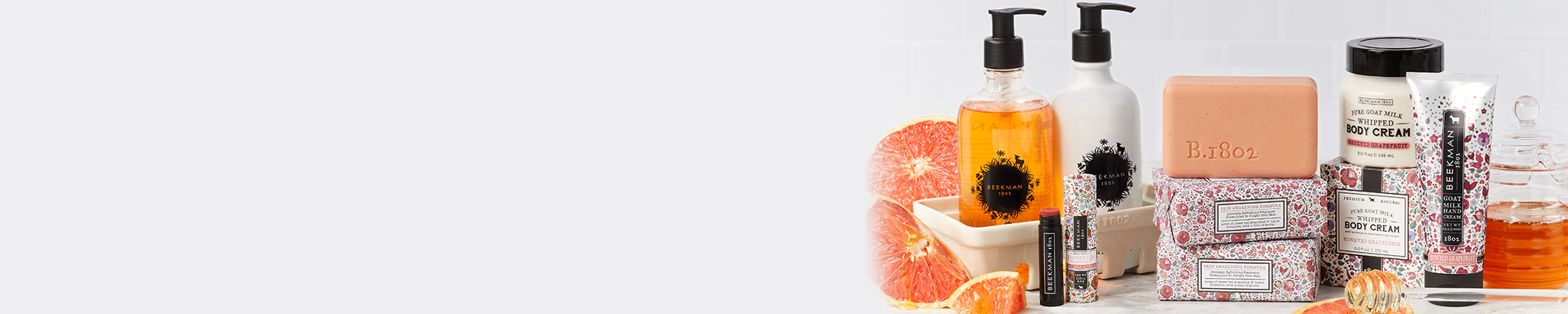 Products from Beekman 1802's Honeyed Grapefruit collection which includes the caddy set, lip balm, bar soap, and body creams to the far right of the screen, surrounded by slices of grapefruit and a honey stick spilling honey on a light grey tile background. 
