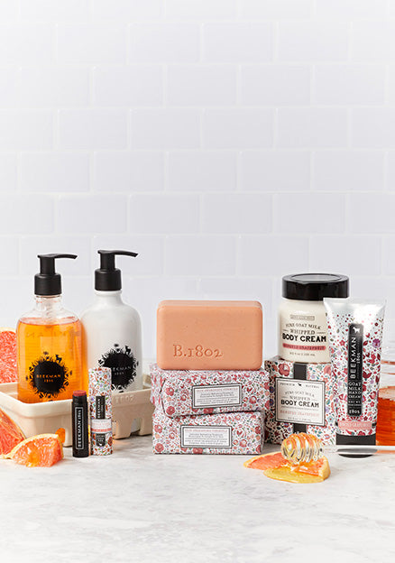 Products from Beekman 1802's Honeyed Grapefruit collection which includes the caddy set, lip balm, bar soap, and body creams, surrounded by slices of grapefruit and a honey stick spilling honey on a light grey tile background. 