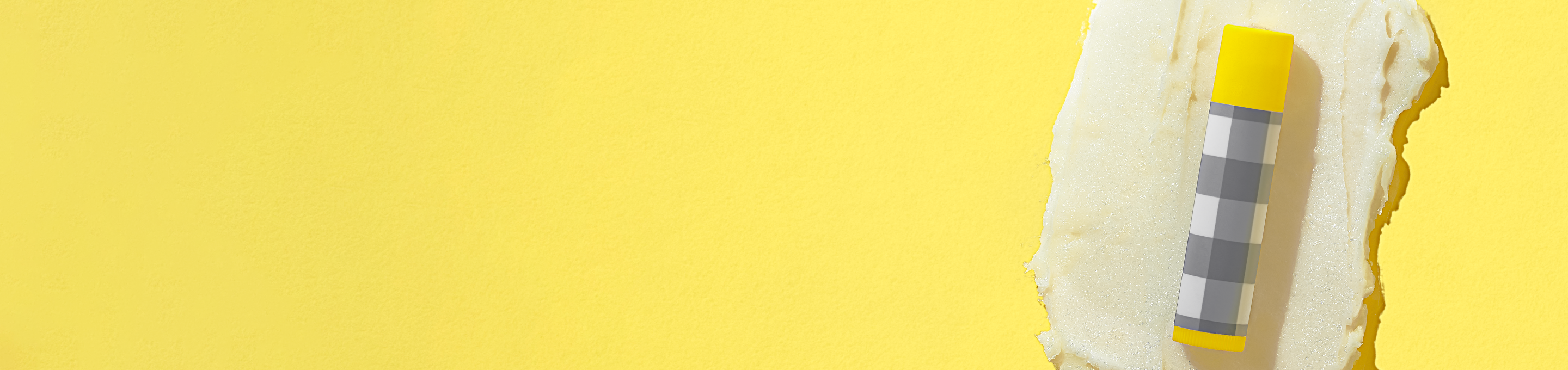 Small Tube of Beekman 1802's Pure Goat Milk Lip Balm in gingham packaging with a yellow cap to the far right of the screen, on top of lip balm ingredient smear on a yellow background. 