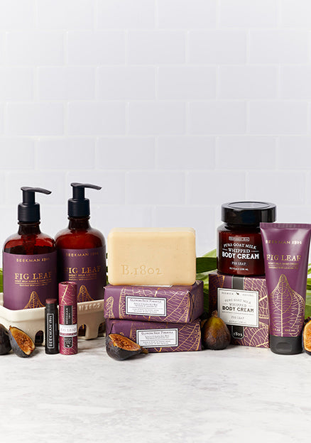 Products from Beekman 1802's Fig Leaf collection which includes the caddy set, lip balm, bar soaps, body cream, and hand cream all displayed next to each other, surrounded by sliced figs and leaves on a light grey tile background. 